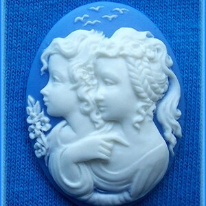 May include: A blue cameo with two white figures of women embracing. The cameo is set against a blue background with white birds flying overhead.