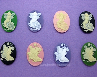 May include: Eight oval-shaped cameo-style resin cabochons with a white fairy design. The fairies are sitting on a flower and have wings. The cabochons are in various colors: green, gray, pink, black, light pink, and blue.