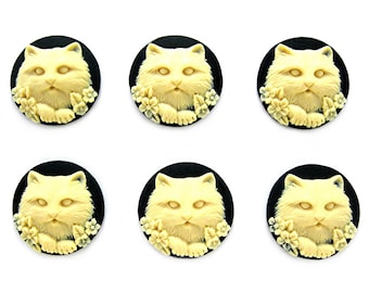 May include: Six cream-colored cat face buttons with black backgrounds. The cat faces are in a 3D design with flowers around the edges.