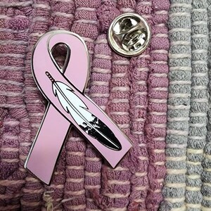 May include: A pink ribbon with a black and white feather design, pinned to a pink and grey fabric background.