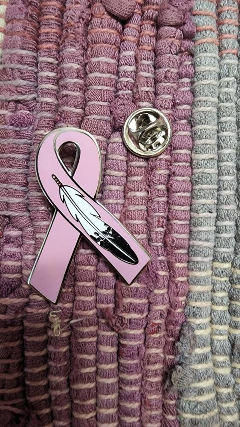 May include: A pink ribbon with a black and white feather design, pinned to a pink and grey fabric background.