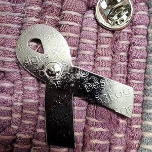 May include: A silver metal ribbon pin with the words "Design is a Process" and "Design is a Process" on it. The pin is on a purple and white striped fabric.