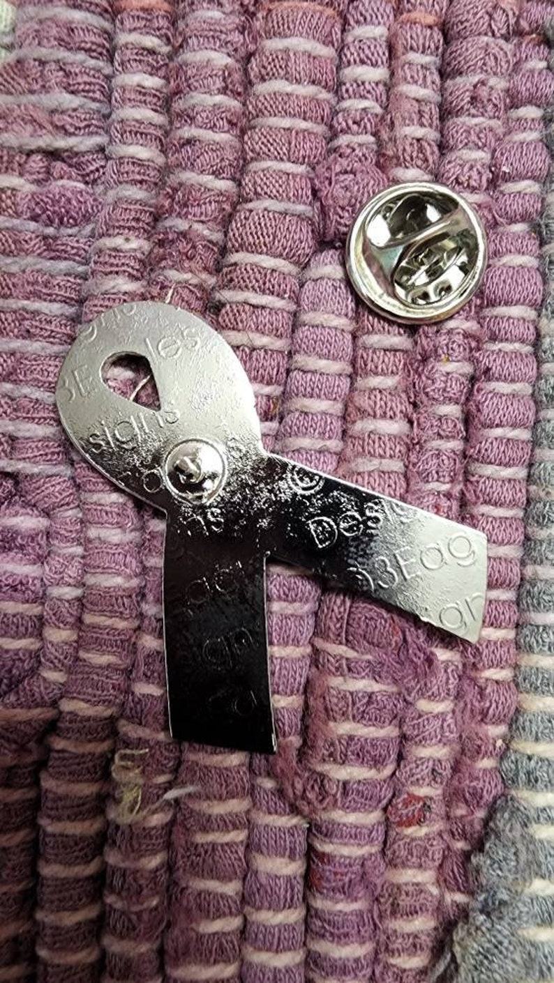 May include: A silver metal ribbon pin with the words "Design is a Process" and "Design is a Process" on it. The pin is on a purple and white striped fabric.