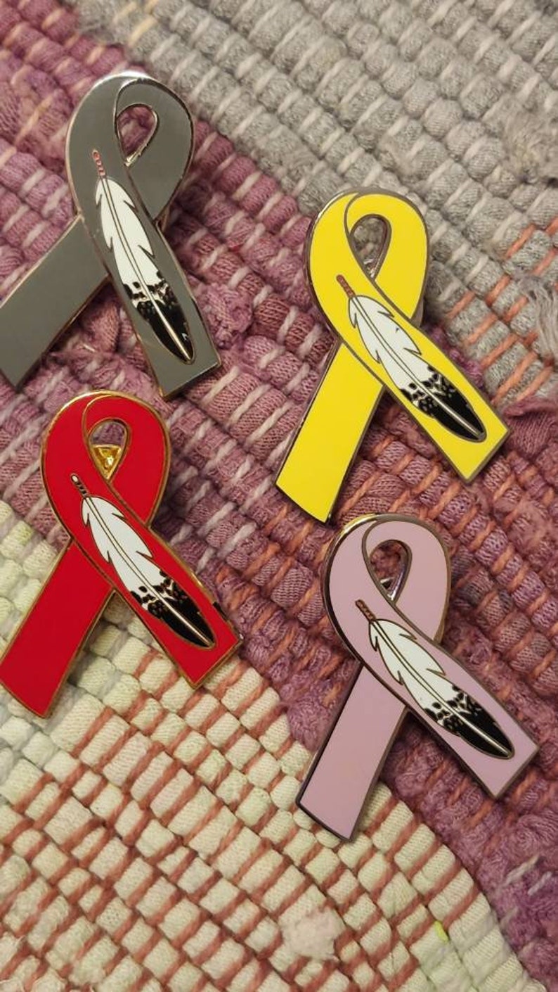 May include: Four enamel pins in the shape of a ribbon with a feather design. The ribbons are gray, yellow, red, and pink.