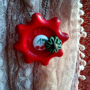 May include: A red, star-shaped pendant necklace with a white cherry design and a green flower charm. The necklace is on a white lace background.