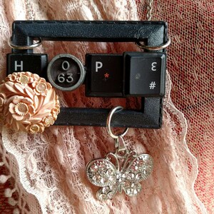 May include: A black frame necklace with typewriter keys spelling "HOPE" and a small, pink, floral brooch. A silver butterfly charm hangs from the bottom of the frame.