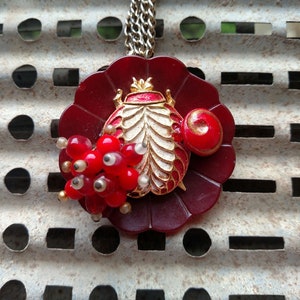 May include: A red and gold pendant necklace featuring a stylized beetle design with a white body and gold accents. The beetle is surrounded by a cluster of red and white beads.