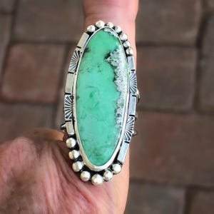 May include: An oval-shaped ring featuring a large, light green stone with natural white and dark inclusions. The stone is set in a silver bezel surrounded by a decorative border of small silver spheres and etched sunburst patterns on the sides.
