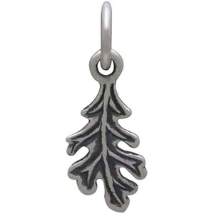 May include: A silver-toned charm in the shape of an oak leaf. The leaf has detailed veining and a textured surface. The charm is suspended from a small, round loop, designed to be attached to a necklace or bracelet. The charm is approximately 1 inch in length.