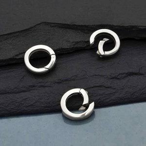 Può includere: Tre chiusure circolari color argento con apertura a cerniera. Le chiusure sono realizzate in metallo lucido e sono disposte su una superficie scura e strutturata. Le chiusure sono progettate per aprirsi e chiudersi, probabilmente per l'uso in gioielleria o accessori.