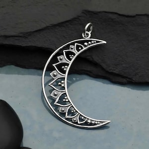 May include: A silver crescent moon pendant with a detailed floral design. The pendant features a series of petal-shaped elements and small, round accents. The jewelry piece has a loop at the top for a chain.