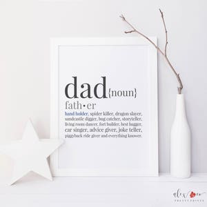 Op de afbeelding: Een ingelijste print met het woord "dad" en de definitie ervan. De definitie bevat zinnen als "hand holder" en "spider killer". Een witte ster en een witte vaas met een tak staan ernaast.