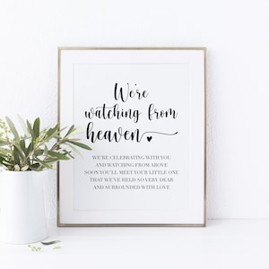 May include: A framed print with the text "We're watching from heaven" in elegant black script. The print includes additional text below, and a small heart symbol. A white vase with green foliage is in the foreground.