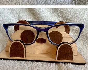 May include: A wooden eyeglasses holder in the shape of a bear. The bear has brown accents for the eyes, nose, and ears. The holder is designed to hold a pair of eyeglasses, as shown in the image with blue-framed glasses.