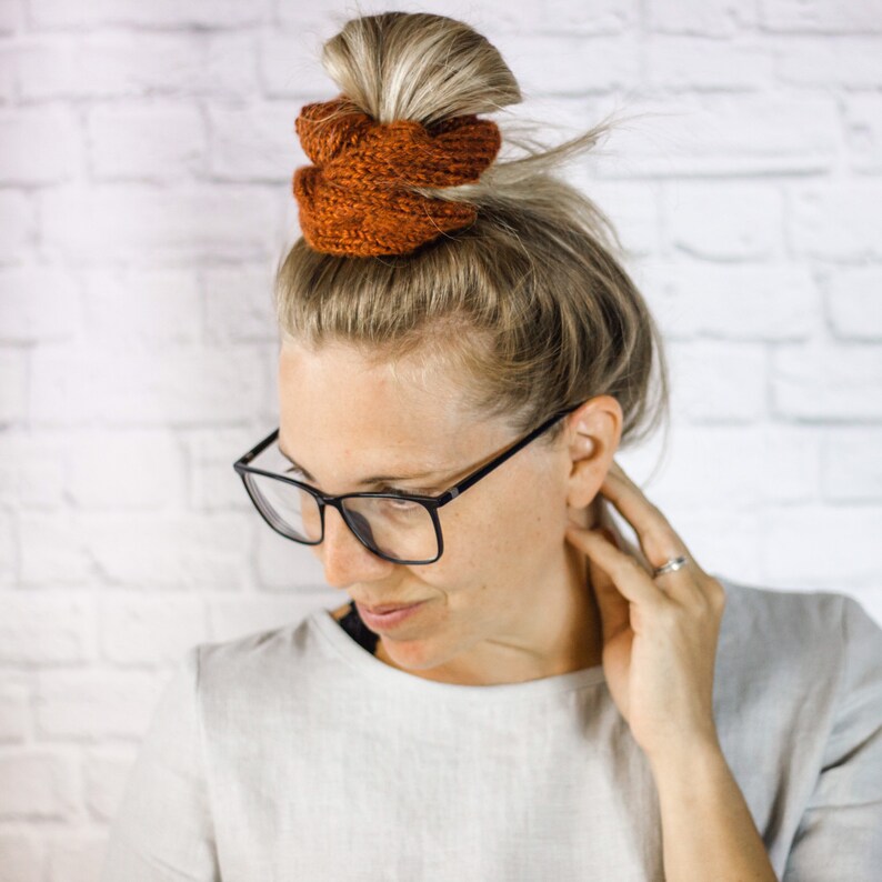 May include: A woman with blonde hair is wearing glasses and a white shirt. Her hair is pulled back into a bun and secured with two brown knitted hair scrunchies.