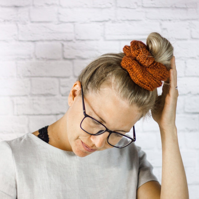 May include: A woman with blonde hair wearing black framed glasses and a brown knitted hair scrunchie.