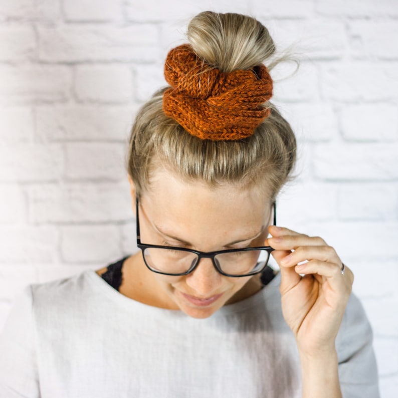 May include: A woman with blonde hair wearing glasses and a brown knitted hair scrunchie.