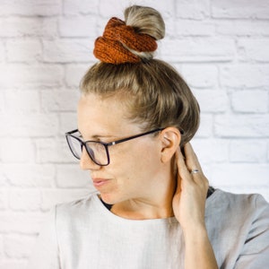 May include: A woman with blonde hair wearing glasses and a brown knitted hair scrunchie. She is wearing a light brown shirt and has her hand on her neck.