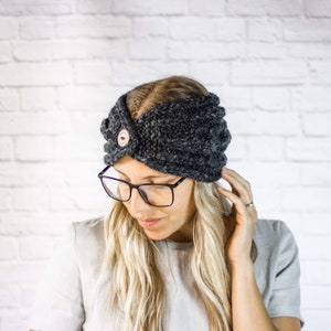 May include: A grey knitted headband with a button detail. The headband is being worn by a person with long blonde hair and glasses.