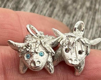 May include: Two silver-toned Highland cow charms. Each charm features a detailed cow head with horns, a loop for hanging, and gemstone eyes. One cow has blue eyes, the other has brown eyes. The charms are displayed against a blurred background.