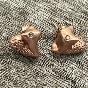 May include: A pair of copper-colored fox-shaped stud earrings. Each earring features a textured body and a small, round gemstone. The fox's head is angled upwards, with a pointed ear and a small gemstone eye.