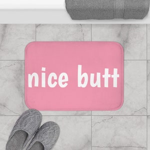 Puede incluir: Una alfombra de baño rectangular rosa con las palabras "nice butt" en letras blancas. La alfombra está sobre un suelo embaldosado, con un par de zapatillas grises y una toalla gris visible.
