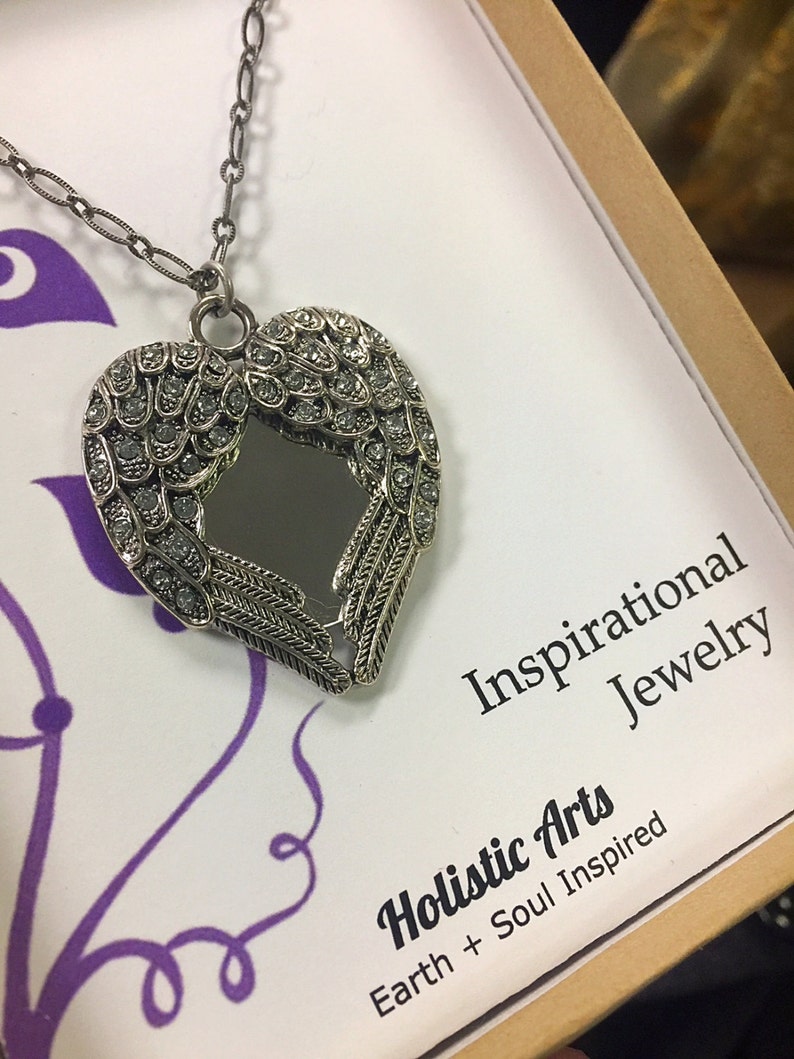 May include: Silver-toned heart-shaped pendant necklace with crystal accents and a mirrored center. The pendant features wing-like designs and hangs on a delicate chain. The packaging reads "Inspirational Jewelry" and "Holistic Arts Earth + Soul Inspired."