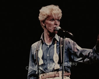 May include: A vintage photograph of a performer on stage. The performer is wearing a blue button-down shirt, cream-colored pants, and suspenders. The performer is holding a microphone and singing.
