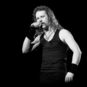 May include: Black and white image of a person singing into a microphone. The person is wearing a black tank top, wristbands, and a necklace. The background is solid black.