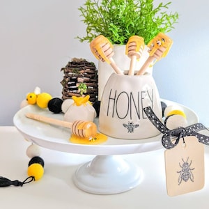 Puede incluir: Un tarro de miel de cerámica blanca con la palabra "HONEY" y un gráfico de abeja, que contiene cucharas de miel de madera. Una exhibición decorativa con temática de abejas incluye un soporte para pasteles blanco, una guirnalda de bolas de fieltro y una etiqueta con una ilustración de abeja.