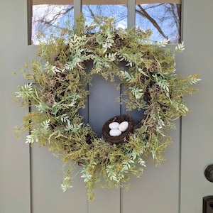 May include: A green and white spring wreath with a bird's nest and three white eggs in the centre.