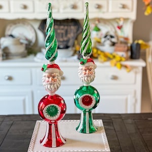 May include: Two Christmas tree toppers featuring Santa Claus heads, red and green spheres, and spiraled green tops. One has a red base, the other green. The toppers are on a white tray, set against a blurred background of a white cabinet.