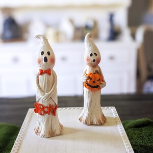 Op de afbeelding: Twee Halloween spookbeeldjes. Eén spook draagt een rode vlinderdas en houdt een bord met "Spooky". Het andere spook houdt een oranje pompoen vast. De spoken zijn wit met perzikwangen en zwarte ogen.