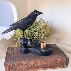 Pode incluir: Estatueta de corvo de madeira preta pousada sobre uma base decorativa com um pequeno castiçal cor de cobre. O corvo está sobre uma base preta, sentado em um livro vintage. Uma pequena planta está atrás do corvo.