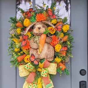 Puede incluir: Una corona de primavera con un conejito marrón que sostiene una zanahoria, rodeado de flores amarillas y naranjas, hojas verdes y mariposas naranjas. Un lazo amarillo y verde con lunares blancos cuelga debajo del conejito.