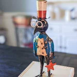 May include: A whimsical metal sculpture of a bird-like figure wearing a top hat, spectacles, and a blue jacket. The figure holds a red star and stands on a decorative base. The hat is red, white, and silver.