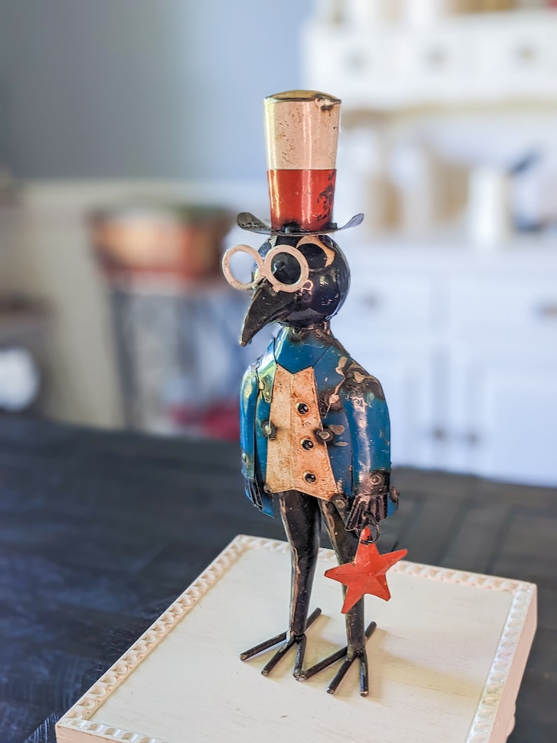 May include: A whimsical metal sculpture of a bird-like figure wearing a top hat, spectacles, and a blue jacket. The figure holds a red star and stands on a decorative base. The hat is red, white, and silver.