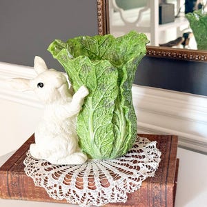 Könnte beinhalten: Eine weiße Hasenfigur lehnt an einer grünen, kohlförmigen Vase. Die Vase und der Hase stehen auf einem alten Buch mit einer Spitzendecke. Die Szene spielt vor einem neutralen Hintergrund und erzeugt eine charmante, dekorative Darstellung.