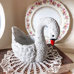 May include: A white swan-shaped planter with a textured surface, black eyes, and a red beak. The planter sits on a vintage book atop a white lace doily. A decorative plate with floral patterns is in the background.