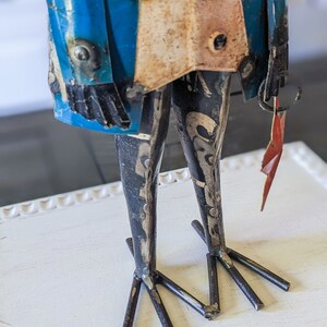 May include: A metal sculpture of a figure with a blue coat, a cream-colored vest, and black legs. The figure holds a small red flag. The sculpture has a distressed, vintage appearance and is set against a white background.