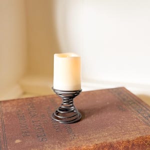 May include: A white candle sits on a black metal spring-shaped candle holder. The candle holder is on a brown book with gold lettering.