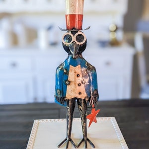 May include: A metal bird sculpture in a patriotic theme. The bird wears a top hat with red, white, and blue stripes, a blue jacket, and holds a red star. The sculpture has a whimsical, folk art style.
