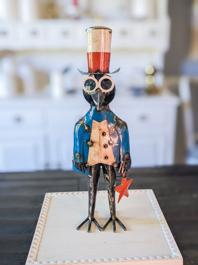 May include: A metal bird sculpture in a patriotic theme. The bird wears a top hat with red, white, and blue stripes, a blue jacket, and holds a red star. The sculpture has a whimsical, folk art style.