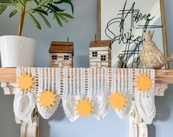 May include: A white crocheted lace runner draped over a mantel, featuring a hanging garland of five yellow sun-shaped wooden cutouts strung together with twine. The sun cutouts are flat, circular, and have radiating points around their edges.