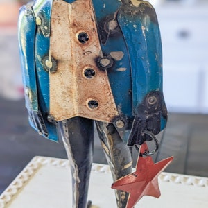 May include: A metal frog figurine wearing a blue jacket with a cream-colored vest and black buttons. The frog holds a red star ornament. The figurine has black legs and feet and is on a white surface.