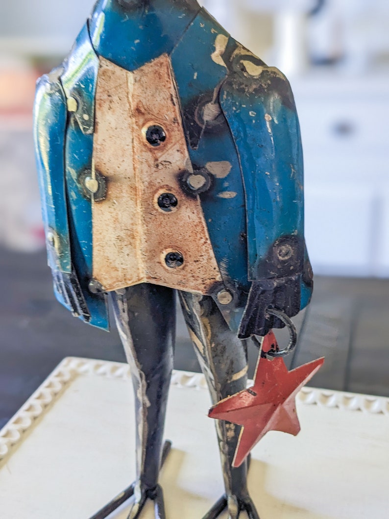 May include: A metal frog figurine wearing a blue jacket with a cream-colored vest and black buttons. The frog holds a red star ornament. The figurine has black legs and feet and is on a white surface.