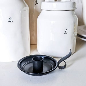 May include: A black metal candlestick holder with a round base and a handle. The holder is in front of several white ceramic jars with black numbers printed on them. The jars are of varying sizes and shapes.