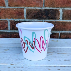 Puede incluir: Cubo de metal blanco con borde enrollado, decorado con corazones superpuestos en verde azulado, burdeos y rosa. El cubo es cilíndrico y se asienta sobre una superficie de madera blanca, con una pared de ladrillo al fondo.