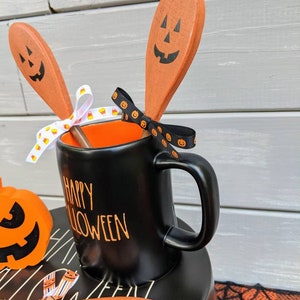 Op de afbeelding: Zwarte mok met oranje tekst "Happy Halloween", gevuld met drie oranje houten lepels met zwarte pompoengezichten. De lepels zijn vastgebonden met een zwart-wit lint met pompoengezichten.