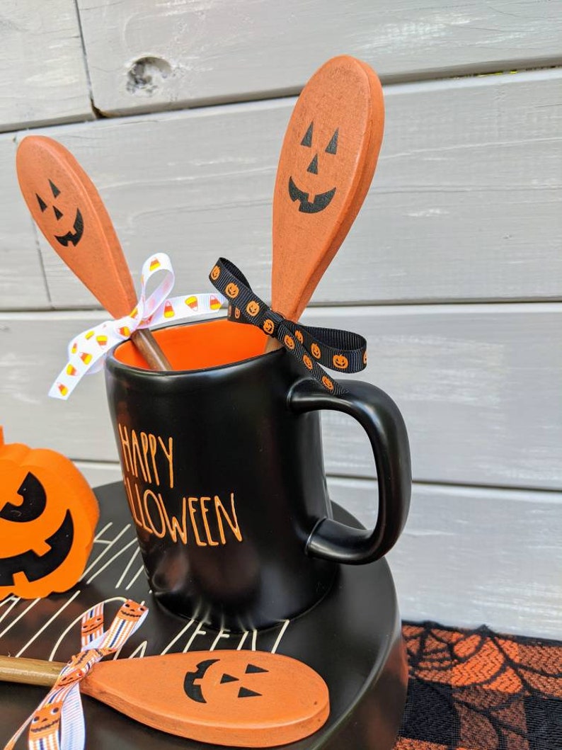 Op de afbeelding: Zwarte mok met oranje tekst "Happy Halloween", gevuld met drie oranje houten lepels met zwarte pompoengezichten. De lepels zijn vastgebonden met een zwart-wit lint met pompoengezichten.